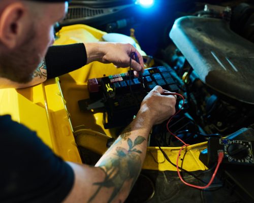 Mechanic of maintenance service carrying out technical diagnostics of car engine and other parts with help of digital multimeter